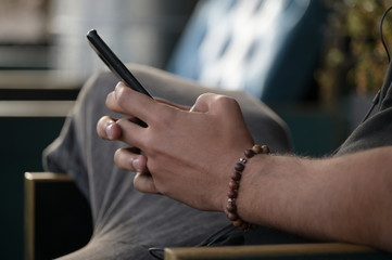 A young spanish man with trendy jewellery at his arm and fingers sits on a wooden chair while he is writing messages on his smartphone.