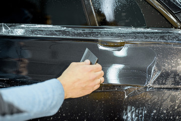 Worker sticking anti-gravel film on a car body with scrapper at the detailing vehicle workshop, close-up. Concept of car body protection with special films