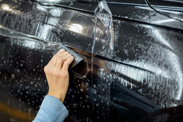 Fototapeta na wymiar Worker sticking anti-gravel film on a car body with scrapper at the detailing vehicle workshop, close-up. Concept of car body protection with special films