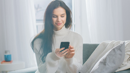 Young Woman Using Smartphone while Sitting on the Chair. Sensual Girl Wearing Sweater Surfs...
