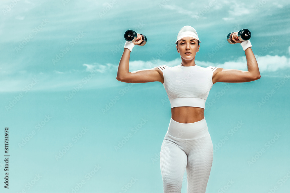 Wall mural Sporty and fit woman athlete doing training with dumbbells on the sky background. The concept of a healthy lifestyle and sport. Individual sports recreation.