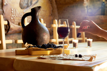 Woman hand with mulberry, glass of red wine, clay jug on wooden table in a wine cellar restaurant