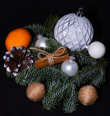 Art installation on a black background for New Year or Christmas from spruce twigs, glass balls, cord balls, pine cone, one mandarin and cinnamon sticks.