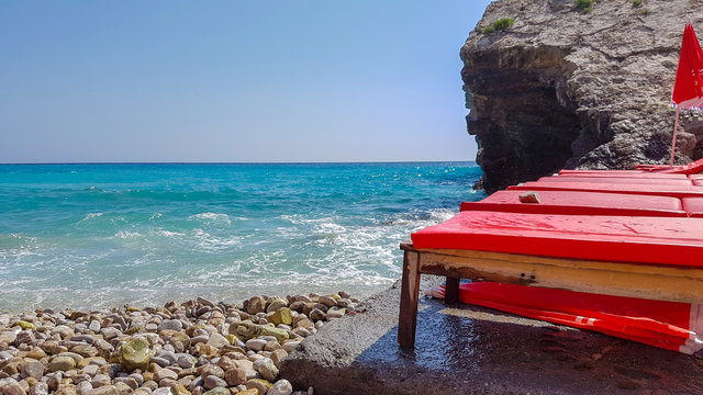 Panoramic view of pebble beach with clear azure blue water, layered rocks and red beach umbrellas, beautiful mediterranean Adriatic Sea coast, Montenegro, selective focus