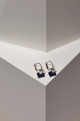 Subject shot of a pair of hoop earrings on the white surface in the space with geometric design. Each earring is decorated with a gem flower with crystals and a blue gem in steel square setting. 