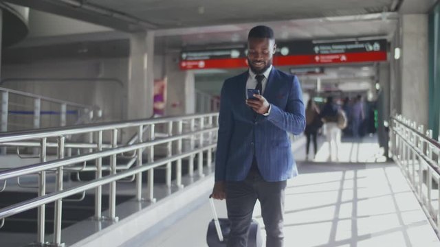 Portrait of young afro entrepreneur using smartphone while walking with luggage in airport