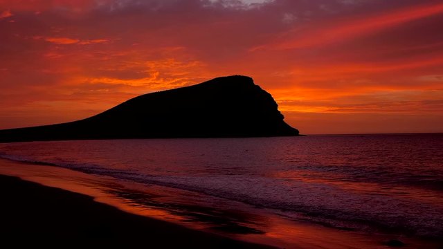 Wide angle shot of an amazing red sunrise at a beach and the silhouette of an old volcano under a cloudy sky. HD cropped edit