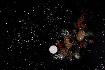Forest pinecones, red berries and green leaves with white candle on the black background in snow.