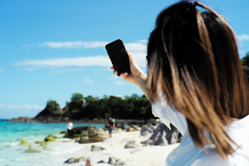 Fototapeta na wymiar Back portrait of female tourist holds mobile phone and takes selfie picture with blurred seascape and people in background