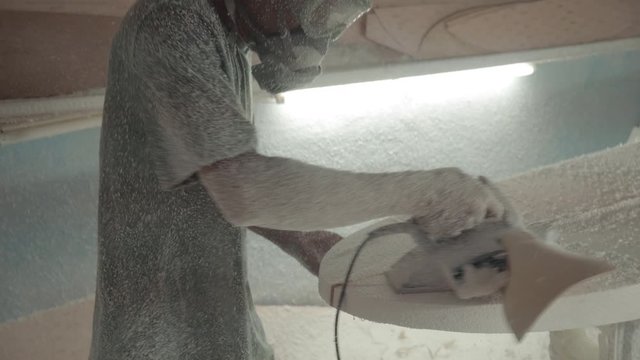 Sequence of shots of Indonesian worker wearing respirator, goggles and protective headphones sanding surfboard lying on workbench using electrical grinding machine