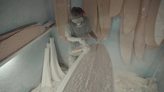 High angle view of Indonesian craftsman wearing safety mask, goggles and headphones sanding surfboard using electric polishing machine