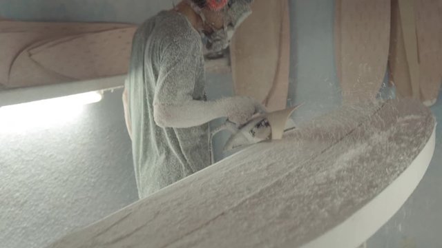 Medium shot of Indonesian craftsman wearing mask, safety goggles and headphones polishing surfboard using automatic polishing machine