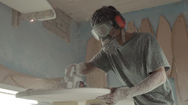 Lockdown of professional surfboard shaper wearing protective equipment polishing surfboard in workshop