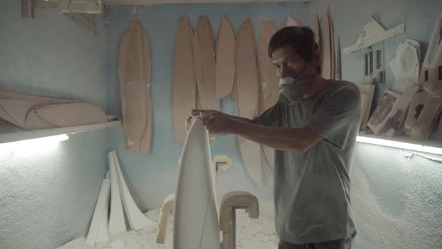 Medium shot of professional surfboard shaper wearing protective mask polishing edges of surfboard using piece of abrasive paper