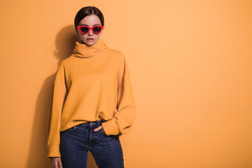 Full size picture of young model stand alone and pose. Wear yellow sweater and dark pants. Stylish red sunglasses. Isolated over yellow background.