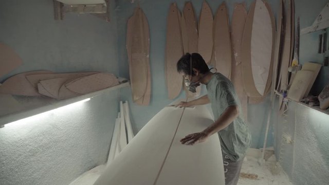 Sequence of shots of Indonesian man blowing off sawdust from surfboard then polishing it with sandpaper, putting off respirator, looking at camera and going away
