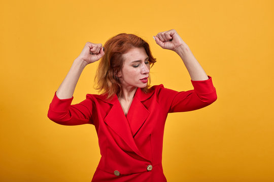 Cheerful caucasian young woman keeping fists up, dancing, enjoying wearing fashion red jacket over isolated orange background. People lifestyle concept.