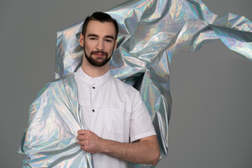 A guy in white clothes. epic stands wrapped iridescent holographic fabric flying behind a cloak