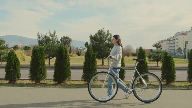 A young and stylish woman walks with a classic pesto bike along the sidewalk near beautiful bushes. traking shot.