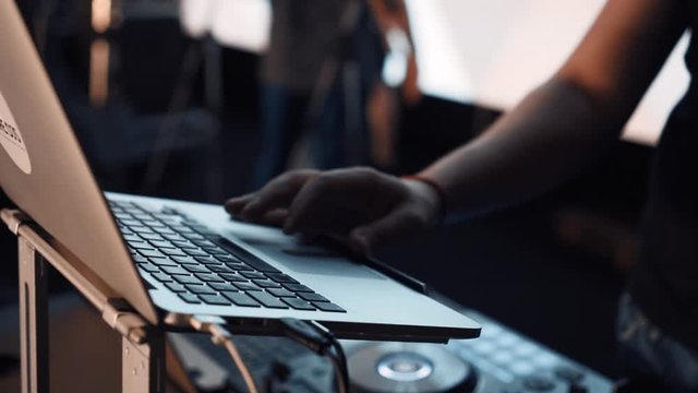 Footage of a professional DJ remote control before a concert on stage. DJ holds his hand on his laptop, setting up equipment at a nightclub party. Disc jockey works.