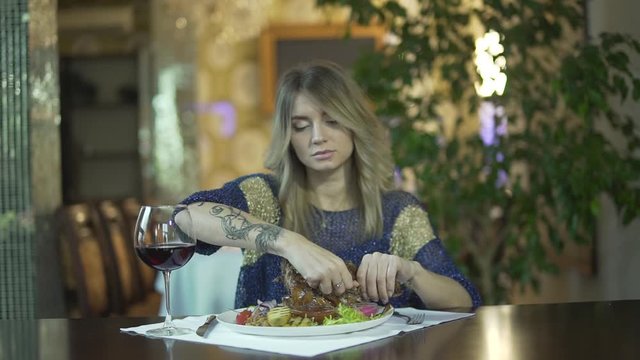 Young beautiful tattooed blond girl in fancy dinner restaurant tearing meat eating with fingers inappropriate behaviour