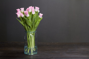 Glass vase with pink tulips on a dark wooden table, greeting background or concept