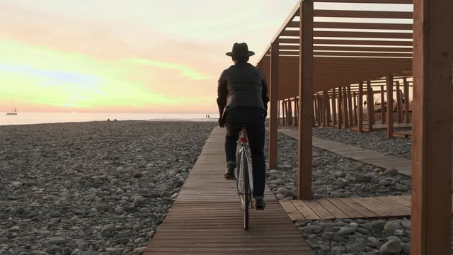 A man in a hat and a leather jacket rides a Bicycle along the embankment near a wooden construction. A man rides a Bicycle on the beach at sunset. Rear view.