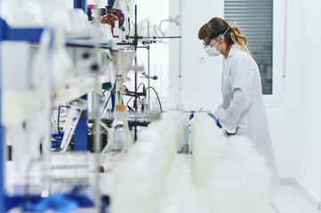 young woman working in the lab. She's wearing a mask, goggles and safety gloves, and she's looking...