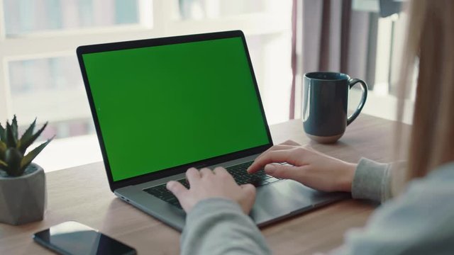 Over shoulder shot of caucasian woman in a grey sweater hands using laptop with green screen in a comfortable, light room. Modern devices, social networks, freelancer. Programming. Slow motion, close