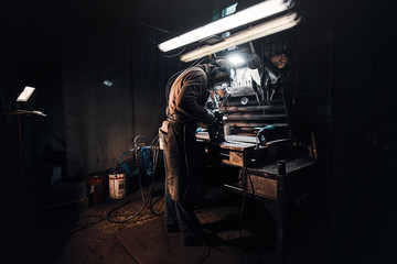 At the dark workshop experienced worker in protective uniform is working with metal.