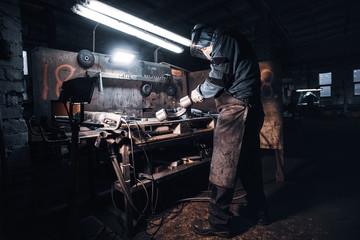At the dark workshop experienced worker in protective uniform is working with metal.