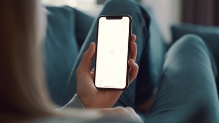 Girl with blond hair lying on blue couch and holding smartphone with green screen in her hand at light modern room. Relaxed woman looking at the mobile phone. - Powered by Adobe
