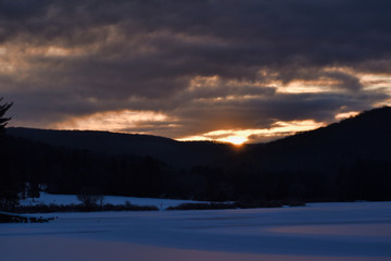 Fototapeta na wymiar Cold Sunrise Over Tree Covered Hills And Frozen Water With Snow At Red House Lake, Allegany State Park, New York