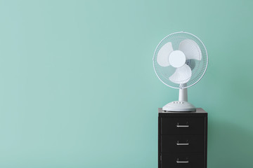 Electric fan on chest of drawers near color wall - obrazy, fototapety, plakaty
