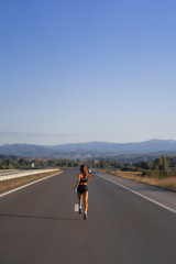 Young girl running along the road. Athletic happy woman jogging in trendy black sexy top and shorts enjoying the sun exercising. Healthy lifestyle. Perfect fitness body shapes and tan skin