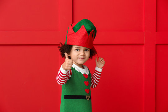 Little girl in costume of elf showing thumb-up gesture on color background