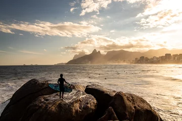 Gordijnen Surfer met surfplank in de hand kijkend naar zonsondergang in Pedra do Arpoador, Arpoador Beach, Rio de Janeiro, Brazilië © Marco Aurelio