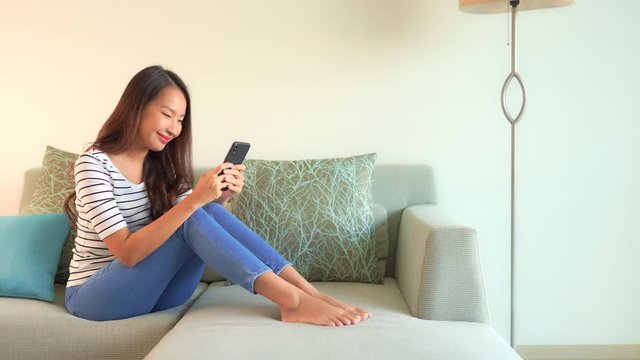 Happy Asian Female Sitting on Sofa and Typing Text Messages on Smartphone. Social Networks Concept With Copy Space