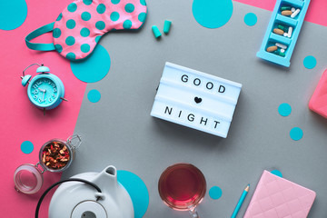 Healthy night sleep creative concept. Sleeping mask, alarm clock, earphones, earplugs and pills. Split two tone, pink and grey paper background with circles and tea. Lightboard with text "Good night".