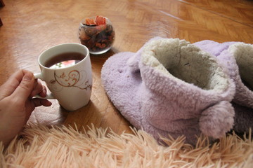 hand holds a mug of tea. slippers on the floor stand and next to the fluffy plaid