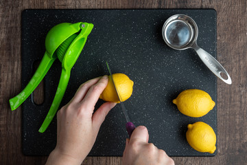 Woman’s hands cutting a lemon on a black cutting board, green citrus squeezer, measuring cup