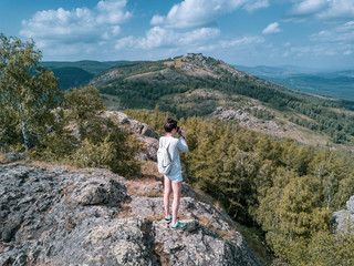Naklejka na ściany i meble Young brunette girl stands on mountain top , takes photo of beautiful Ural landscapes; rocky cliff in summer sunny day