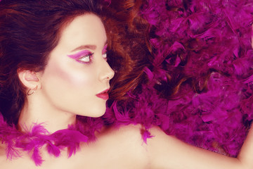 Beautiful young brunette lies with closed eyes in pink feathers. Girl with pink / purple make-up. Beauty.