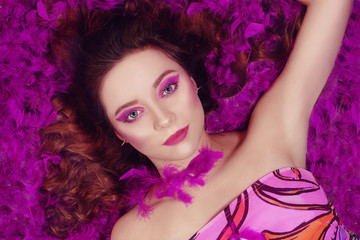 A young girl with purple make-up is looking at the camera. A girl with long locks of hair is looking at the camera. The model lies on purple feathers.