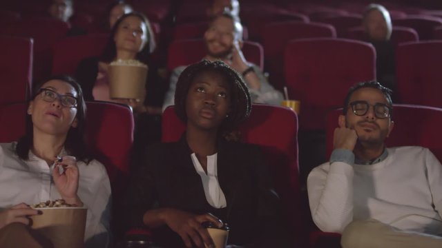 Tracking shot of multi-ethnic people sitting in front row at movie cinema and enjoying film. They are looking at screen and shaking their heads in disapproval, then laughing at joke