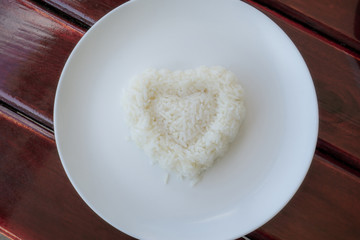 White rice cooked in heart shape in a white plate on brown wooden table, top view photo