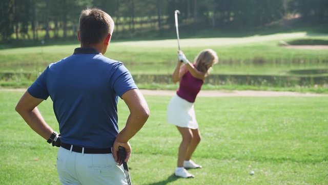 Green park, man trainer teaches woman to play Golf, woman hits the ball, golf course in forest area, green lawn, slow motion.