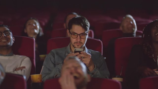 Tracking of bearded man in glasses sitting in movie theater and using mobile phone, then looking up at screen and laughing with audience