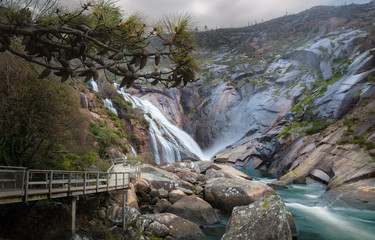 The waterfall near the city of Ezaro in northern Spain in Galicia on the Atlantic coast. It's cloudy. On the left is a path made of wooden planks for pedestrians.