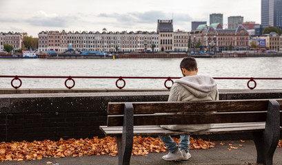 Man rear view sitting on a park bench alone, Rotterdam, Netherlands skyline, cloudy autumn day